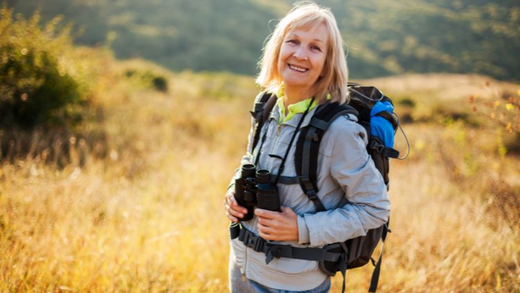 A senior female traveler with binoculars and backpack standing in a field